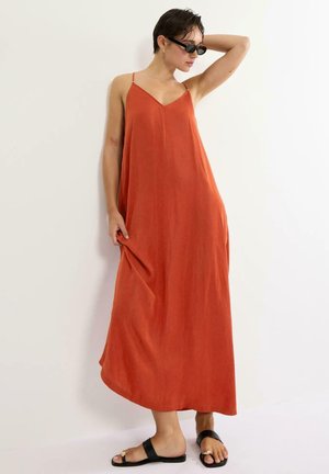 Kobieta pozująca w bezrękawowej długiej sukience w kolorze spalonym pomarańczowym, czarnych okularach przeciwsłonecznych i czarnych płaskich sandałach na białym tle.