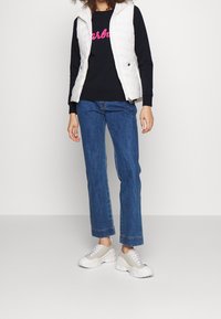 Gilet matelassé blanc par-dessus un sweat à capuche noir avec un texte rose, associé à un jean droit bleu et des baskets gris clair.