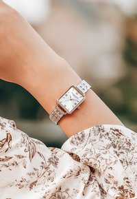Reloj de acero inoxidable plateado con una esfera blanca rectangular, acentos en oro rosa y marcadores de hora en números romanos, llevado en una muñeca.