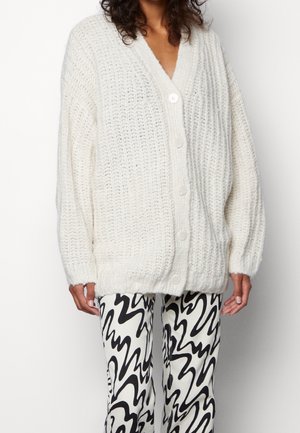 Femme portant un cardigan blanc en maille épaisse boutonné et un pantalon à motif abstrait noir et blanc sur fond uni.