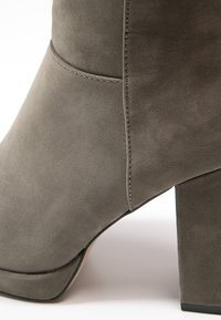 Botte de cheville en suédine grise avec un talon épais, présentant une texture lisse et un design minimaliste. Les coutures latérales sont visibles près du talon.