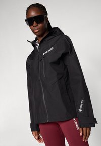 Schwarze, wasserdichte Terrex-Jacke mit Kapuze, ausgestattet mit einem Frontreißverschluss, Seitentaschen und Gore-Tex-Branding. Glatte Textur, modernes Design.
