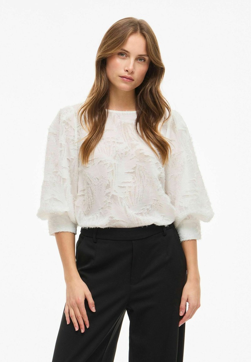 Blouse blanche texturée avec des manches trois-quarts et un col rond, associée à un pantalon noir ajusté. La texture présente un motif subtil.