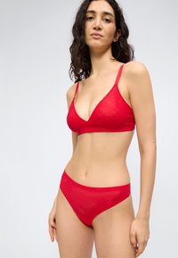 Femme aux cheveux longs et foncés portant un bralette en dentelle rouge et une culotte en dentelle rouge assortie, debout devant un fond clair uni.