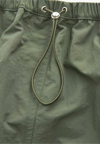 Grüne Stoffshorts mit elastischem Bund und einer durchgezogenen Schnur, die mit einem runden metallischen Stopper gesichert ist.