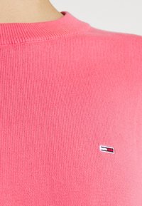 Gros plan d'une personne portant un pull en tricot rose avec un petit logo rectangulaire rouge, blanc et bleu sur la poitrine.