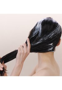 Cheveux noirs et mouillés avec un produit de conditionnement blanc appliqué. Une main tient une mèche de cheveux tandis que l'arrière d'un cou est visible ; arrière-plan neutre.