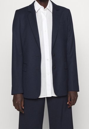 Persoon die een donkerblauw colbert en broek draagt over een wit overhemd met verticale strepen, staande tegen een effen achtergrond.