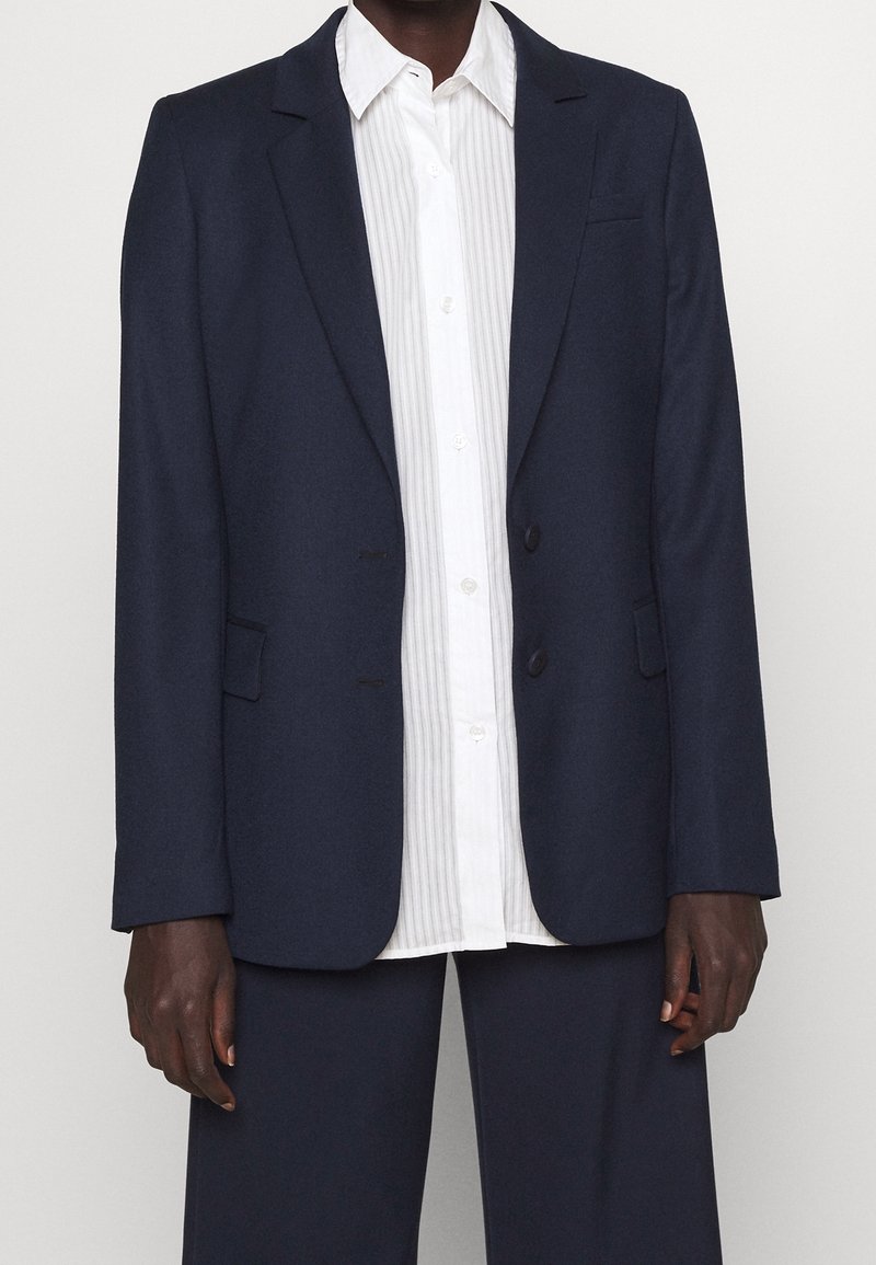 Personne portant une veste et un pantalon de costume bleu marine foncé sur une chemise blanche à rayures verticales, debout devant un fond uni.