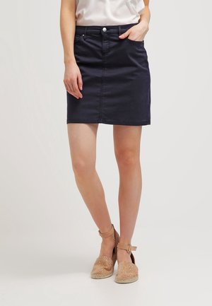 Kvinde iført en mørkeblå knælang nederdel med forlommer, hvid top og beige kilehælssandaler med ankelrem mod en ensfarvet baggrund.