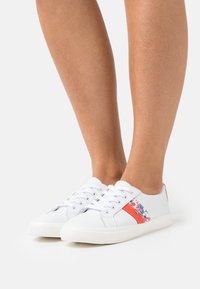 Vita låga sneakers med röd och blå blommig rand, som bärs på barfota mot en vit bakgrund.