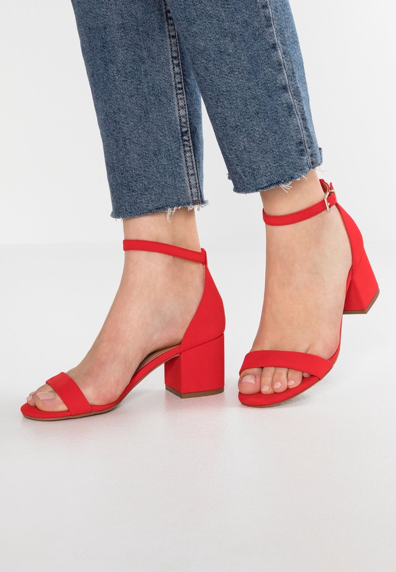Pieds portant des sandales rouges à talon bloc avec brides à la cheville associées à un jean bleu à ourlet effiloché sur un fond blanc uni.