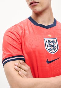 Άτομο που φοράει κόκκινη φανέλα της αγγλικής ποδοσφαιρικής ομάδας με σκούρο μπλε γιακά, έμβλημα με τρία λιοντάρια, λογότυπο Nike και εμφανή σταυρωμένα χέρια.