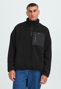 Sudadera de forro polar negra con cuello de media cremallera, tela texturizada y un bolsillo en el pecho con cierre de cremallera. Llevada con pantalones vaqueros de denim azul.
