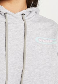 Šedá mikina s kapucí vyrobená z měkké látky s tkaničkou. Na přední části se nachází decentní logo v modré a růžové barvě s nápisem "G-Star RAW DENIM."