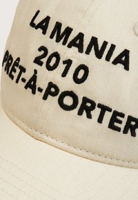 Krém színű baseballsapka ívelt csúccsal és fekete hímzett felirattal: "LA MANIA 2010 PRÊT-À-PORTER" jól láthatóan megjelenítve.