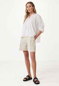 Vit skjorta med knappar och broderade hål detaljer på ärmarna, i kombination med beige plisserade shorts och svarta flätade sandaler.