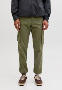 Uomo che indossa pantaloni cargo verde oliva con grandi tasche laterali, sneakers marroni con lacci bianchi e una giacca nera, in piedi davanti a uno sfondo semplice.