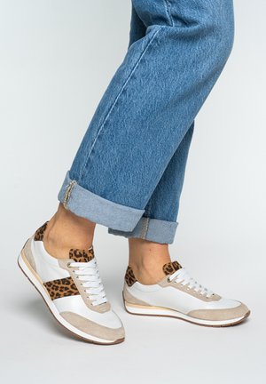 Persoon draagt opgerolde blauwe spijkerbroek en witte sneakers met beige accenten en luipaardprintdetails, staand op een lichtgrijze vloer.