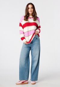 Gebreide trui met rode, roze en crème strepen, gedragen met lichtblauwe broek met wijde pijpen en roze sneakers. Zachte textuur, ontspannen pasvorm.