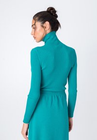Turkizna rebrasta pulover s turtleneck ovratnikom, dolgimi rokavi in tesnim krojem, z visokim ovratnikom in strukturirano tkanino. Prikazan je zadnji pogled.