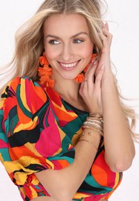 Bunte Bluse mit Puffärmeln in roten, gelben, grünen und schwarzen Mustern; Modell trägt orangefarbene Blumenohrringe und mehrere Armbänder.