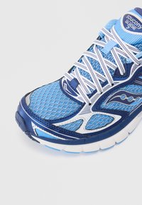 Scarpa da corsa blu con tomaia in mesh, accenti bianchi e blu scuro, superficie strutturata e suola imbottita. Presenta un sistema di allacciatura prominente.