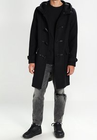 Abrigo de lona negro con capucha, cierres de toggles y bolsillos laterales. Llevado sobre una camisa negra con jeans grises desgastados y zapatillas negras.
