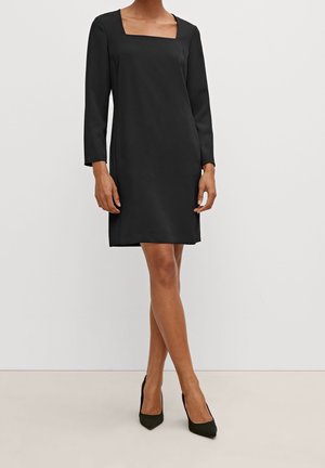 Femme portant une robe noire à manches longues avec un décolleté carré et des talons noirs pointus, debout devant un fond uni.