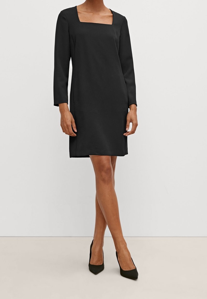 Femme portant une robe noire à manches longues avec un décolleté carré et des talons noirs pointus, debout devant un fond uni.