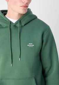Zelená bavlněná mikina s klokaní kapsou, nastavitelnou šňůrkou a bílým textem loga na hrudi. Měkká textura, volný střih.
