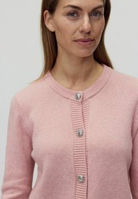 Ljusrosa cardigan gjord av mjukt tyg, med rund halsringning, ribbade detaljer och silverfärgade knappar framtill.