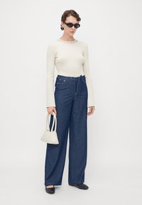 Blue wide-leg denim pants with a high waist and tie detail; paired with a white ribbed long-sleeve top and a small white handbag.