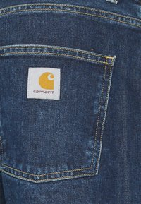 Donkerblauwe denimstof heeft een achterzak met een wit vierkant patch waarop een gele logo staat. Contrasterend stiksel benadrukt de randen van de zak.