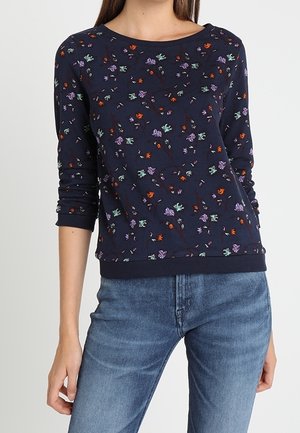 Femme portant un haut à manches longues bleu marine avec de petits motifs floraux colorés et un jean en denim bleu.