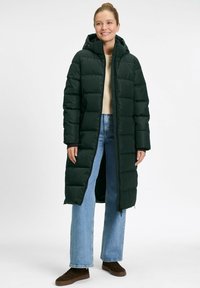 Frau in einem langen dunkelgrünen Daunenmantel über einem beigen Pullover, hellblauen Jeans und braunen Sneakers, steht vor einem schlichten Hintergrund.