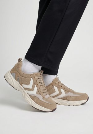 Beige snøresneakers med hvide Chevron-mønstre, båret med hvide sokker og sorte bukser mod en neutral baggrund.