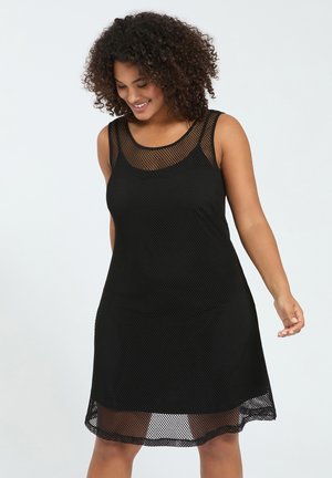 Lächelnde Frau mit lockigem Haar, die ein ärmelloses schwarzes Netzkleid trägt, steht vor einem schlichten hellen Hintergrund.