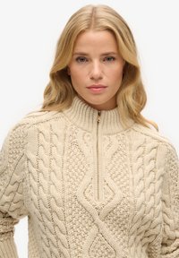 Beige gebreide trui met een hoge geribbelde kraag en een halve ritssluiting, met gemêleerde kabelpatronen aan de voorkant en op de mouwen.