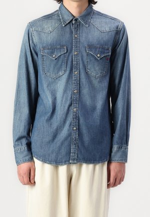 Mężczyzna ubrany w niebieską, dżinsową koszulę na guziki z długim rękawem i dwoma kieszeniami na piersi, w połączeniu z kremowymi spodniami.