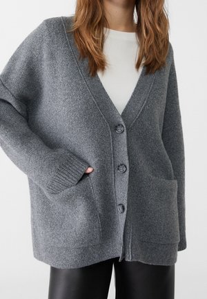 Vrouw draagt een losse grijze gebuttonde cardigan over een wit topje, met één hand in de voorzak, gecombineerd met zwarte broek.