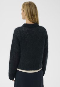 Melnā, teksturētā džemperis ar noapaļotu kakla izgriezumu un garām piedurknēm, saīsinātu apakšu un mīkstu, pūkainu audumu. Nav redzamu rakstu vai metāla detaļu.