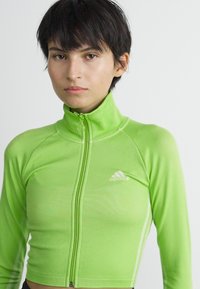 Ungdom med kort mørkt hår iført en limegrønn zip-up Adidas-genser foran en enkel lys bakgrunn.