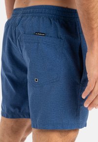 Modré plavkové šortky vyrobené z texturovanej látky, s elastickým pásom a zadnou kapsou s kovovým očkom na odvod vody.