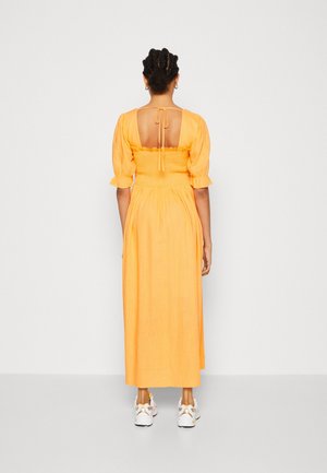 Oranje linnen maxi-jurk met een geplooide lijfje, korte pofmouwen en een lage rug. Gecombineerd met witte sneakers met accenten.