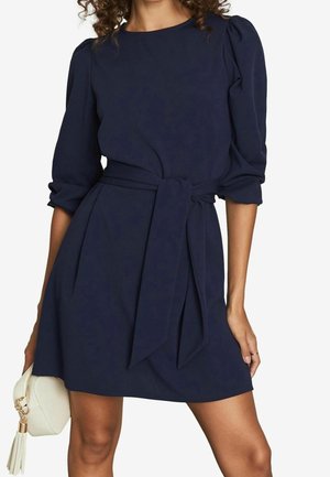 Mujer con un vestido azul marino de manga larga y cinturón, sosteniendo un bolso pequeño blanco con borla.
