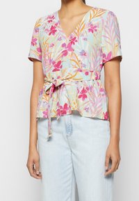 Blouse à manches courtes florale dans des couleurs pastel, avec un décolleté en V, une taille cintrée avec un nœud, et un ourlet en peplum. Tissu doux et léger.
