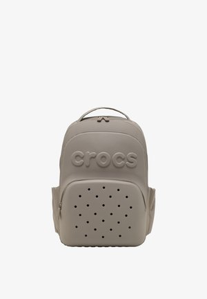 Hellbeige Crocs-Rucksack mit einem oberen Griff, einer Fronttasche mit Reißverschluss und Belüftungsöffnungen sowie dem eingeprägten Crocs-Logo auf der oberen Vorderseite.
