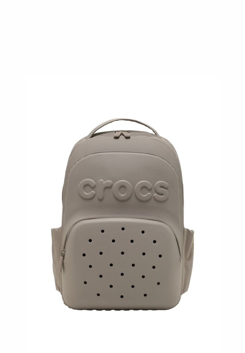 Mochila de Crocs color beige claro con un asa superior, bolsillo frontal con cremallera que cuenta con agujeros de ventilación y logotipo de Crocs en relieve en la parte delantera superior.