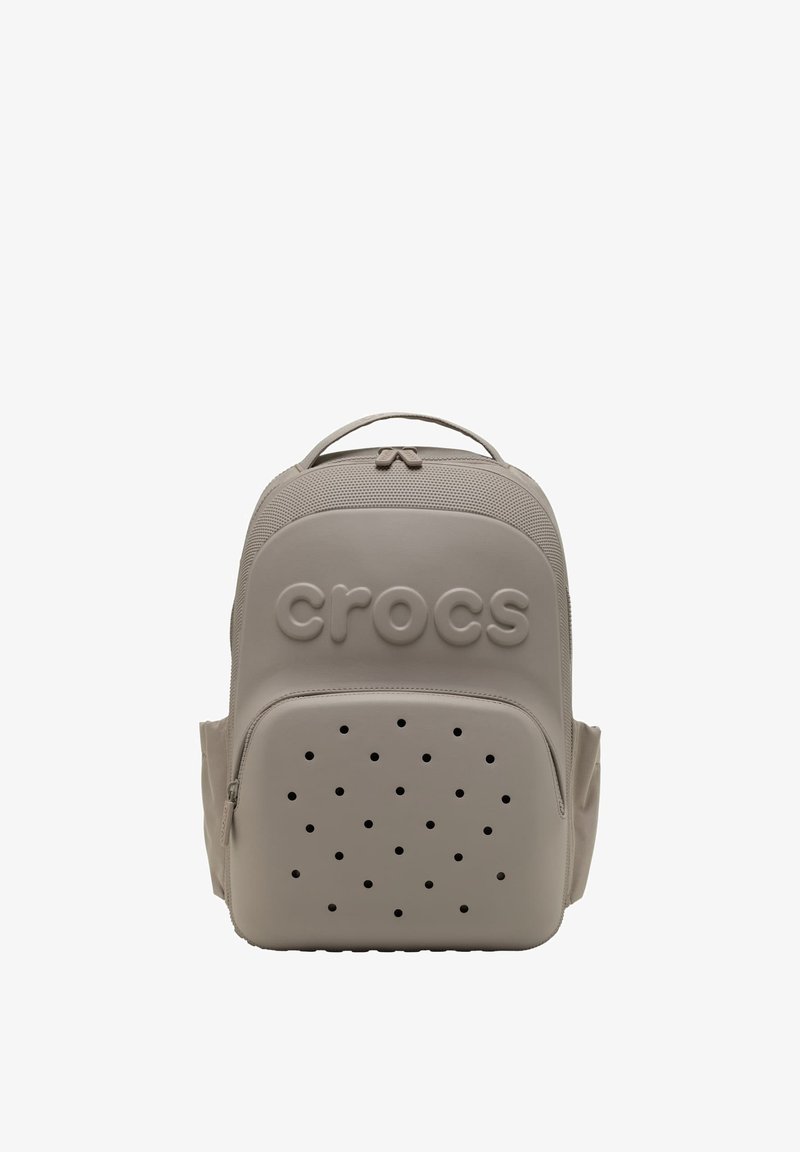 Jasno beżowy plecak Crocs z górnym uchwytem, przednia kieszeń na zamek błyskawiczny z otworami wentylacyjnymi oraz tłoczone logo Crocs na górnej przedniej części.